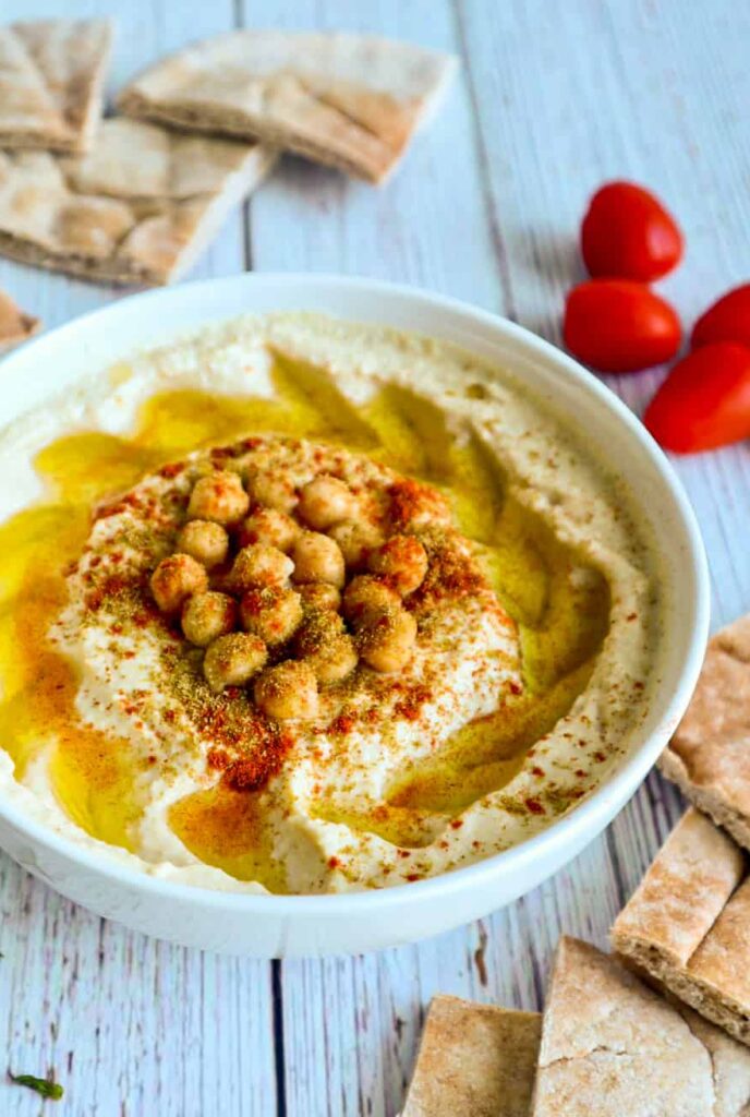A bowl of hummus with chickpeas, olive oil, cumin and paprika on top with slices of pita bread on the side