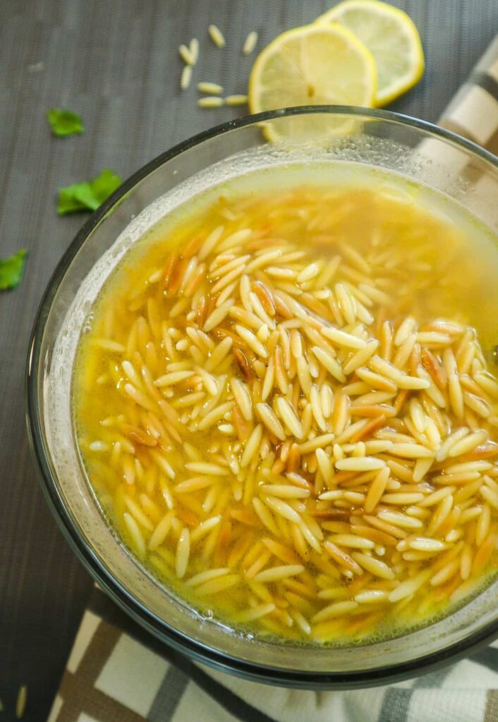 A bowl of Orzo Pasta Soup with 2 slices of lemon next to it.