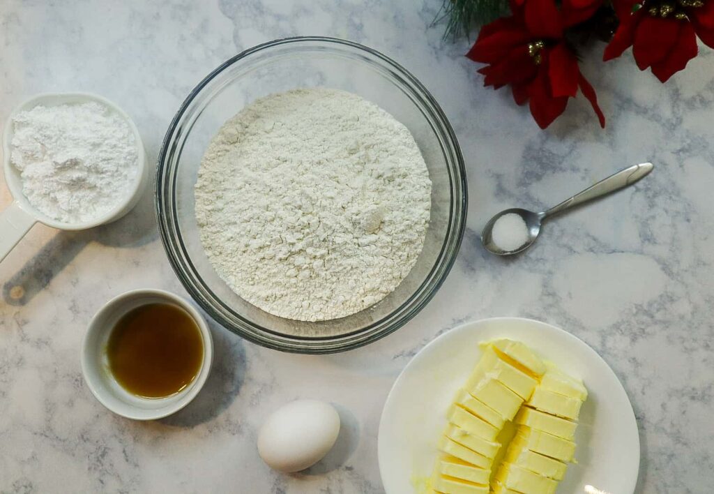 A bowl of flour, an egg, a plate of 2 slices of butter, a cup of vanilla extract, a cup of sugar and a teaspoon of salt
