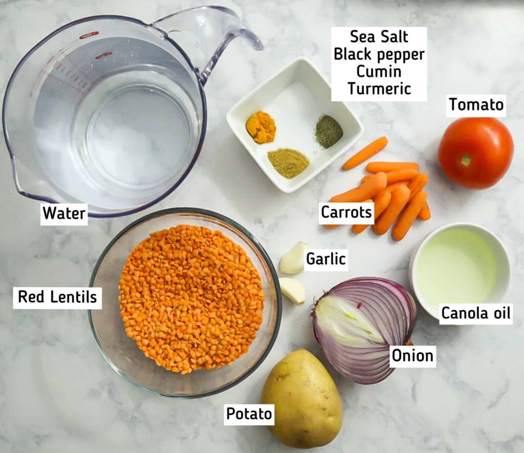A bowl of red lentils, half an onion, 2 cloves of garlic, a tomato, a potato, baby carrots, a bowl of spices, a bowl of canola oil and a measuring cup full of water 