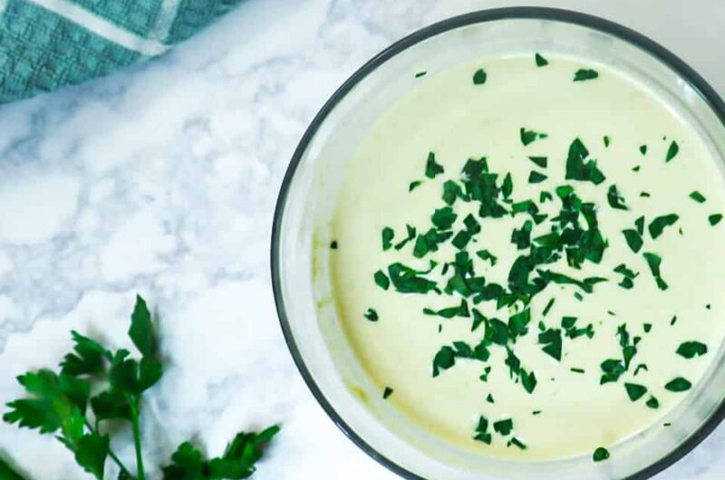 A bowl with Tahini sauce and parsley as a garnish with lemon slices and bunch of parsley.