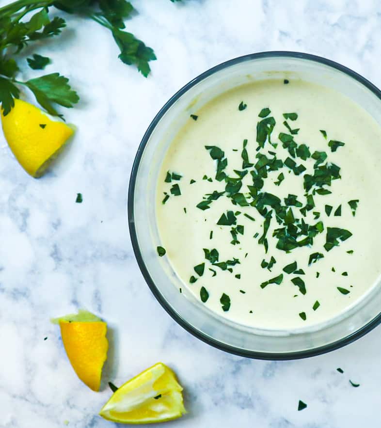 A bowl with Tahini sauce and parsley as a garnish with lemon slices and a bunch of parsley.