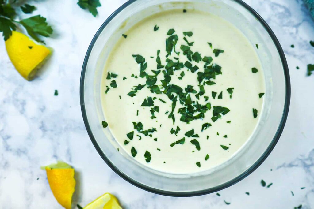 A bowl of tahini sauce and parsley as a garnish with lemon slices and a bunch of parsley. 
