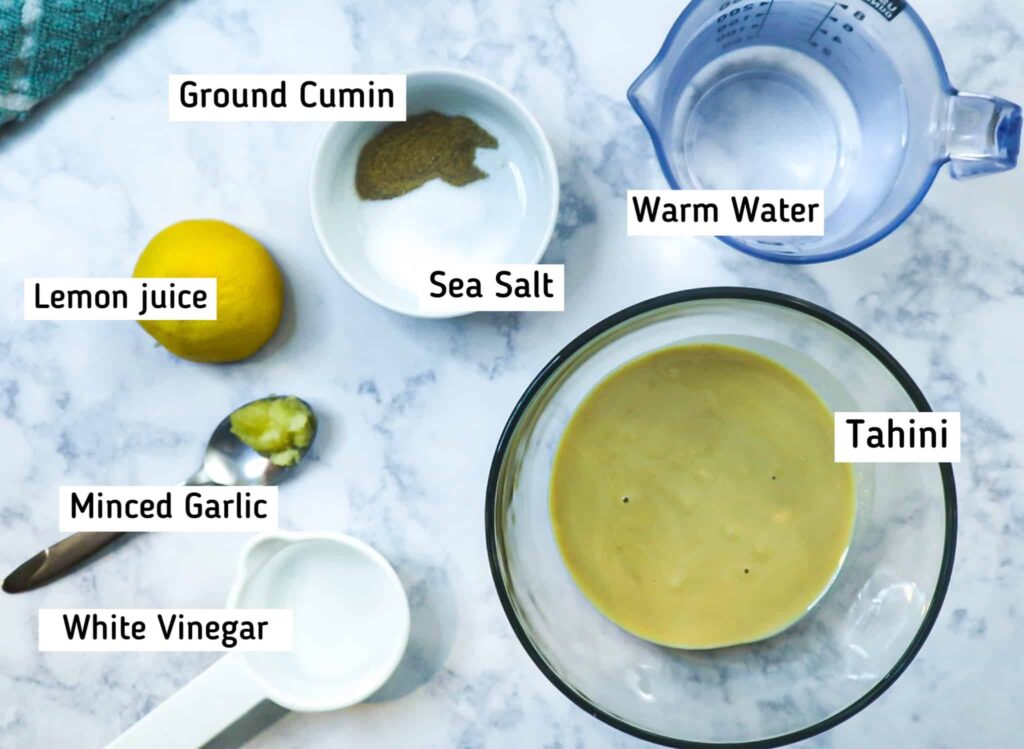 Tahini sauce recipe ingredients are:
A bowl of tahini paste, half a lemon, white vinegar, a spoon with minced garlic. a small bowl with salt and ground cumin and water in a measuring cup 