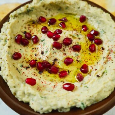 A bowl of a baba ganoush, pomegranate seeds, olive oil, sumac and pieces of pita bread