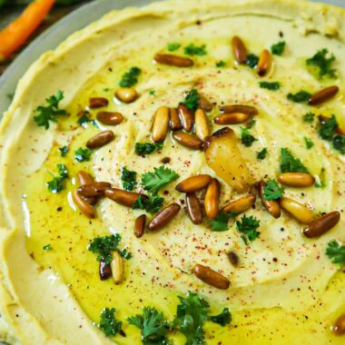 A plate with roasted garlic hummus, pepper slices, pita bread slices, toasted pine nuts, parsley and olive oil