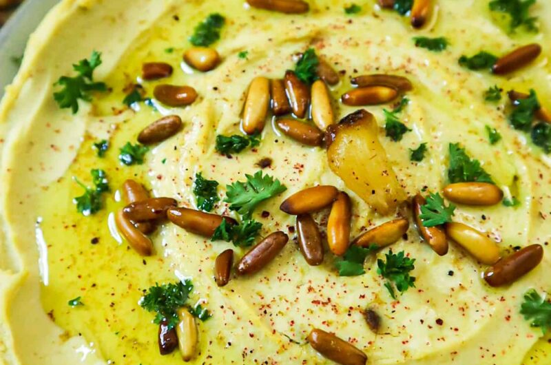 A plate with roasted garlic hummus, pepper slices, pita bread slices, toasted pine nuts, parsley and olive oil