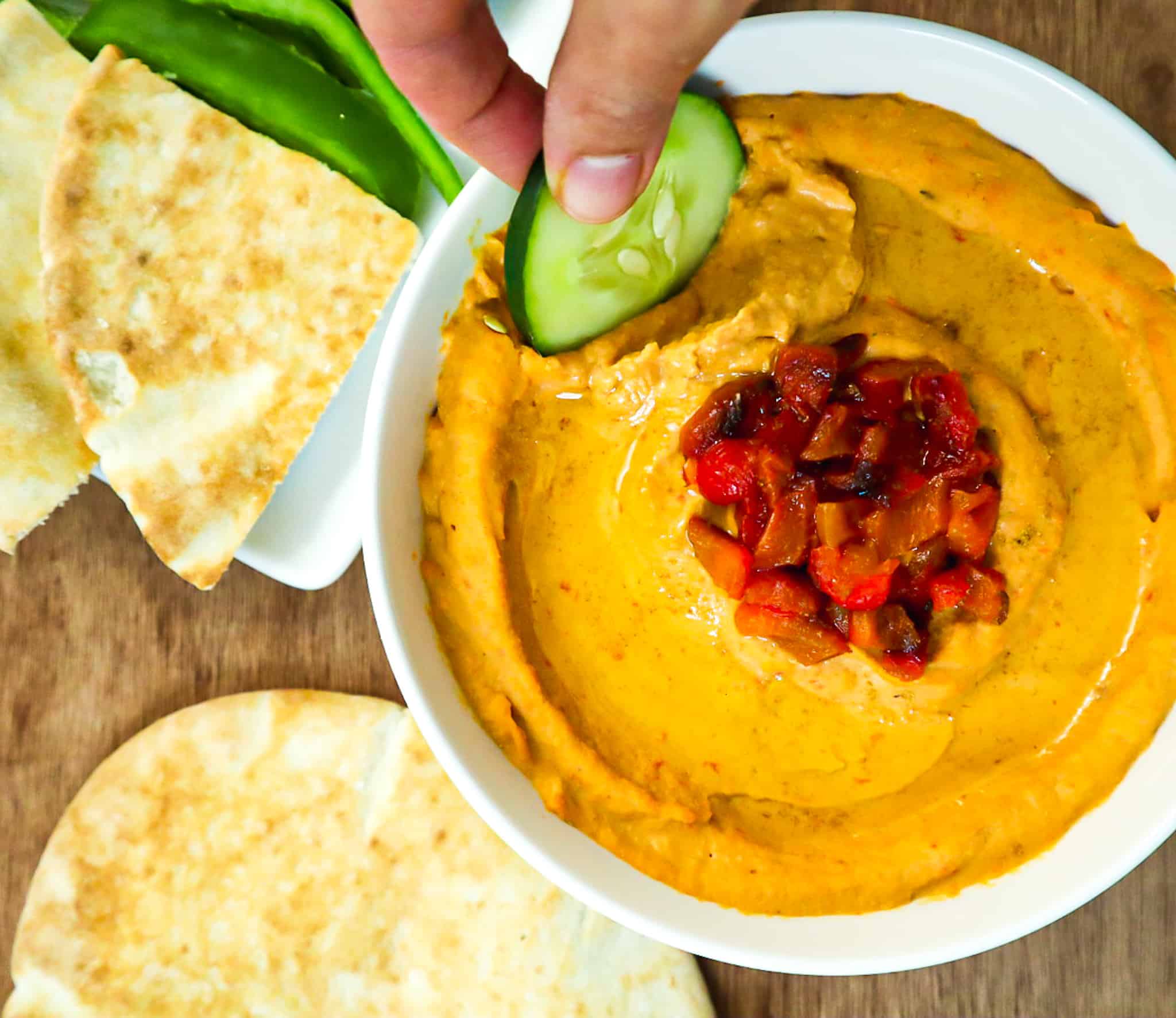 A bowl of roasted red pepper hummus with a cucumber as a dip and pieces of pita bread on the side. 