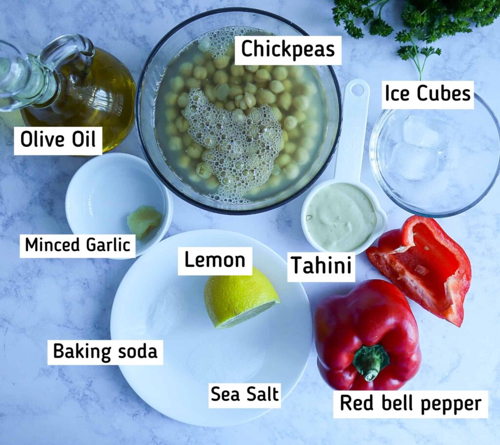 A bowl with chickpeas, a bowl with ice cubes, 1 1/2 red peppers, a cup with tahini, a plate with salt, baking soda and lemon. A bowl with minced garlic and a bottle of olive oil 