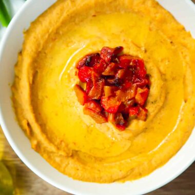 A bowl of Roasted red pepper hummus with diced roasted peppers on top. A plate with sliced pita bread, cucumbers, green and orange peppers.