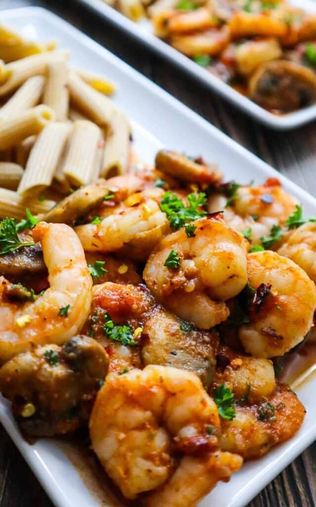 A plate with spicy shrimps and mushrooms with parsley on top and pasta on the side