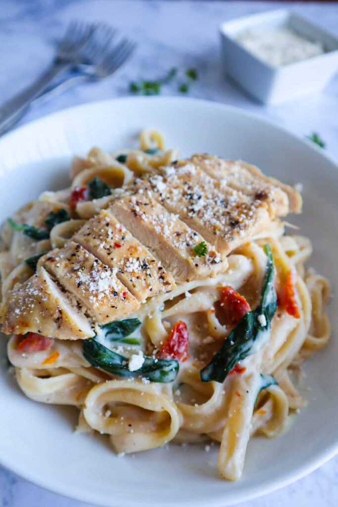 A bowl of fettuccine Alfredo with spinach and sun-dried tomatoes topped with sliced chicken, parmesan and parsley 