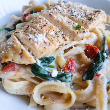 A bowl of fettuccine Alfredo with spinach and sun-dried tomatoes topped with sliced chicken, parmesan and parsley