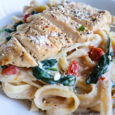 A bowl of fettuccine Alfredo with spinach and sun-dried tomatoes topped with sliced chicken, parmesan and parsley