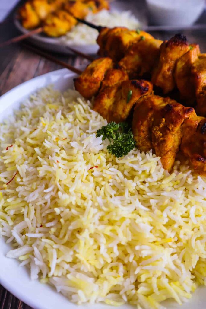 Biryani rice in a dish with cilantro on top, chicken kebabs skewers and raita on the side