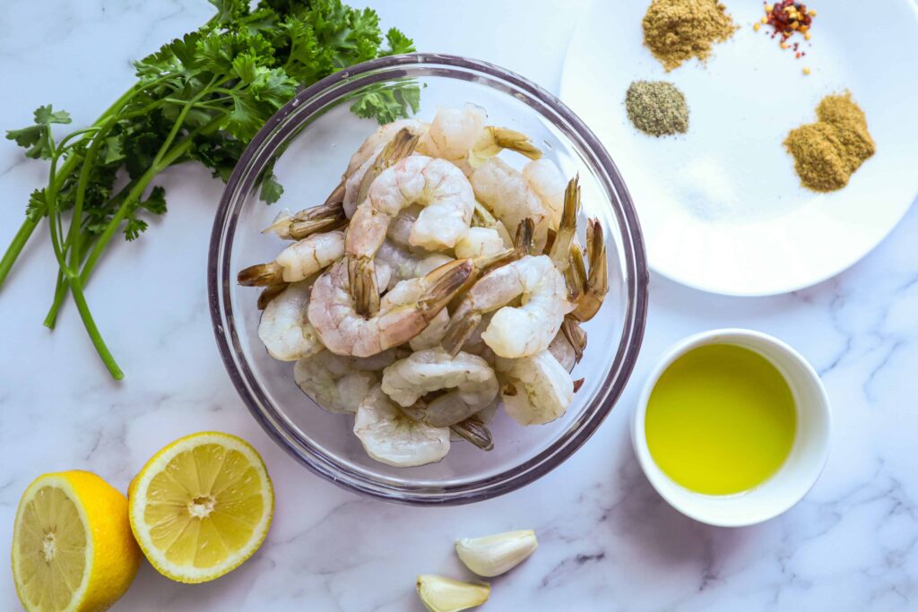 A bowl of raw shrimp, olive oil, spices, lemon and garlic cloves 