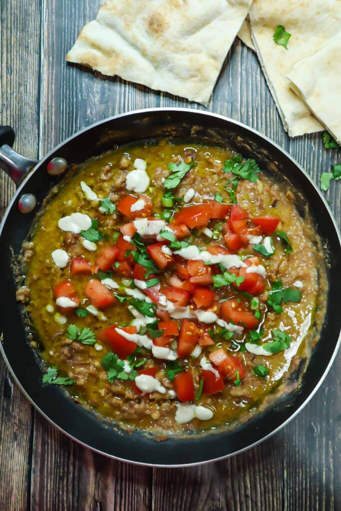 ful medames in a saucepan, topped with diced tomatoes, green onions, cilantro, tahini and olive oil. on the side slices of pita bread