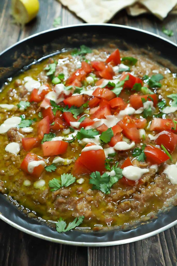 Fava beans in a saucepan, topped with diced tomatoes, green onions, cilantro, tahini and olive oil. on the side slices of pita bread
