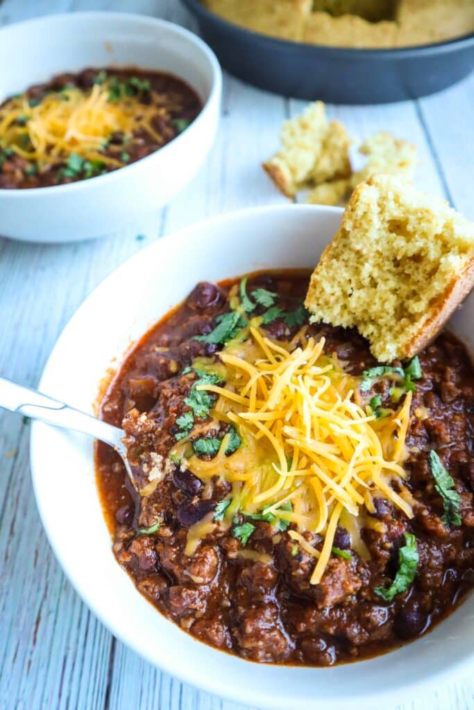 A bowl of chili topped with shredded cheese, chopped cilantro and a piece of a corn bread on the side