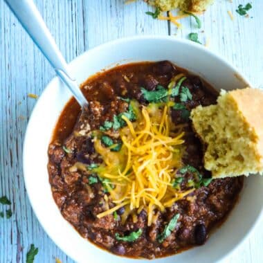 A bowl of chili toped with shredded cheese, chopped cilantro and a piece of a corn bread on the side