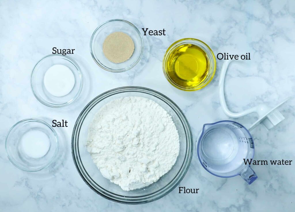 A bowl of flour, paddle attachment, olive oil, water, yeast, sugar and salt