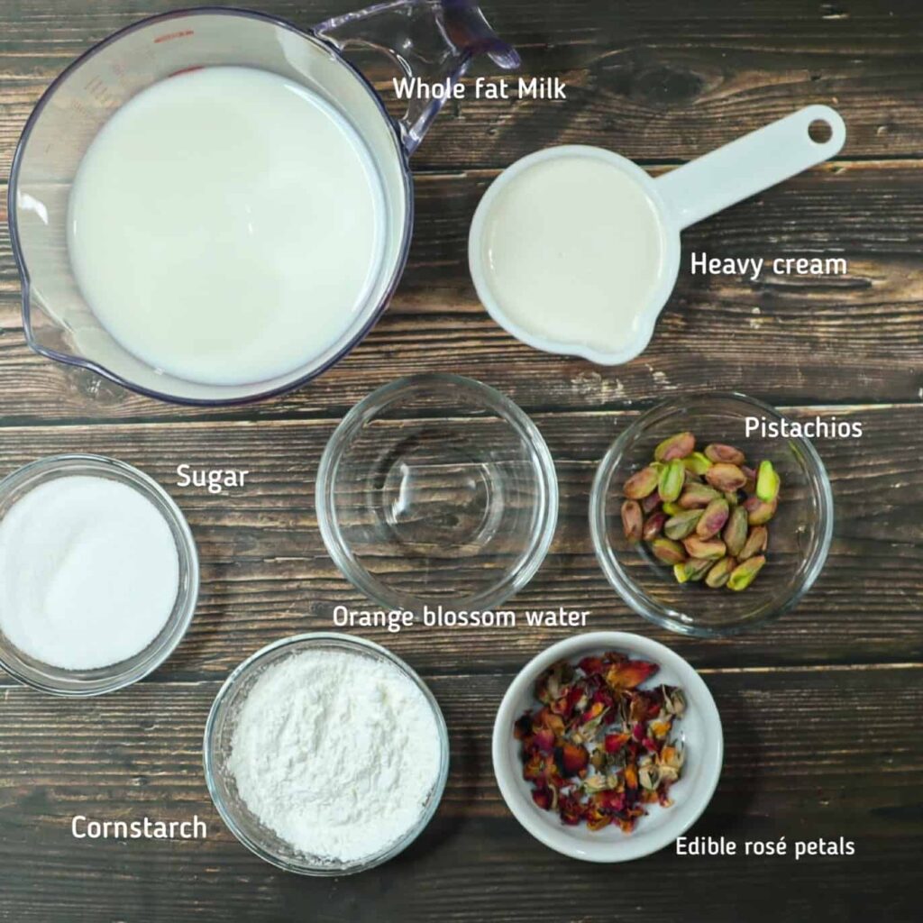 A measuring cup fiiled with milk, heavvy cream, a bowl of sugar, cornstrach, orange blossom, pistachios and rose petals 