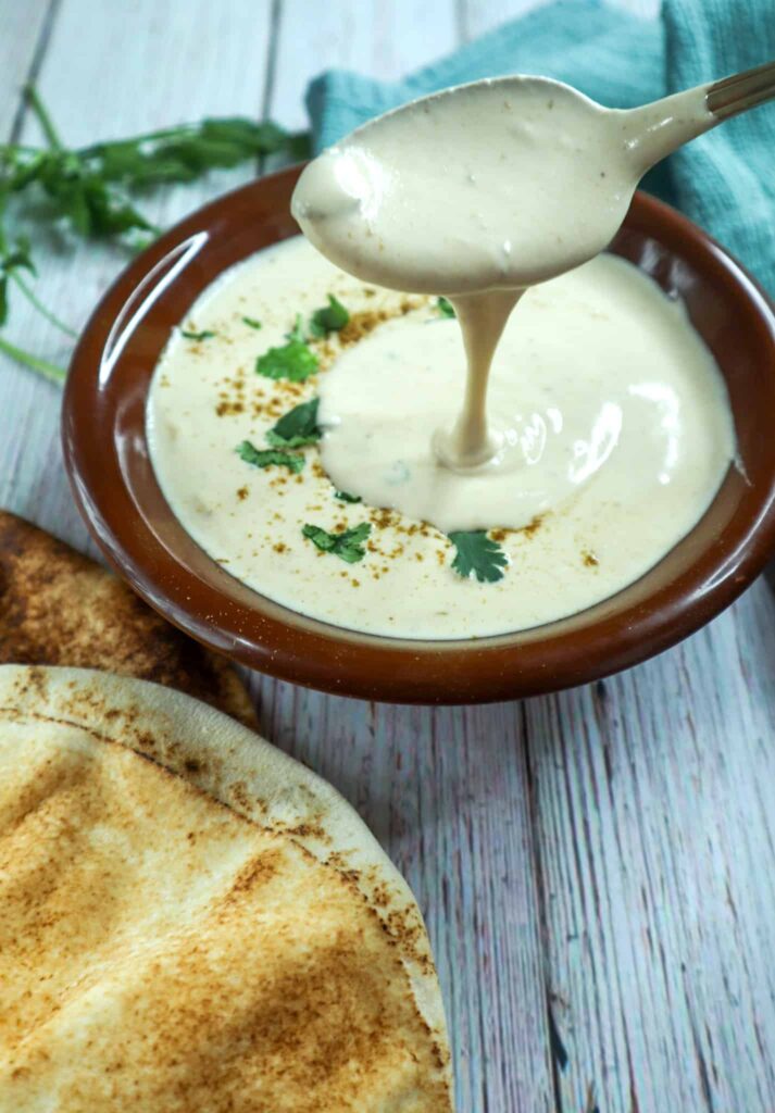 A spoon dripping with the dip with pita bread, parsley and towel on the side
