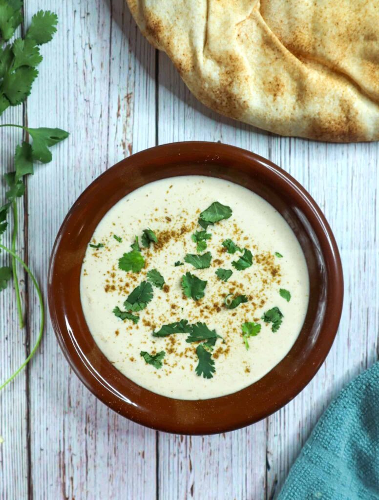 A bowl of Tahini sauce with pita bread, parsley and towel on the side